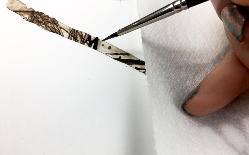 Close up photograph of a hand and fine paintbrush drawing with peat soil. The fingers have chipped nail polish and the artwork is protected by a paper towel.